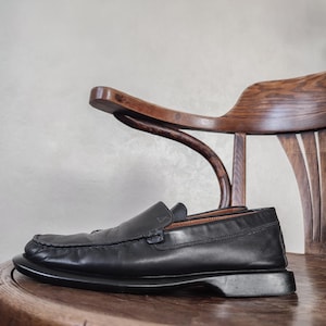 K&ouml;nnte beinhalten: Ein schwarzer Leder-Loafer-Schuh mit runder Spitze, der auf einem Holzstuhl liegt. Der Schuh hat einen niedrigen Absatz und ein klassisches Design. Der Stuhl ist aus dunklem Holz mit geschwungener R&uuml;ckenlehne und Armlehne.