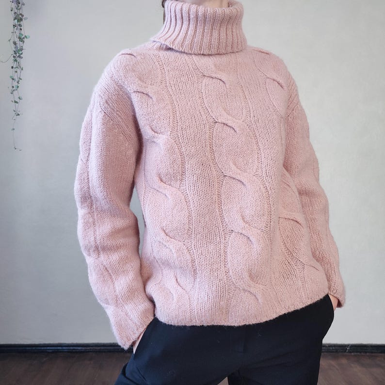 May include: A pale pink cable-knit turtleneck jumper. The jumper has a high, ribbed collar and a textured cable pattern across the front and sleeves. The sleeves are long, and the jumper is made of a soft material. The person is wearing black trousers.