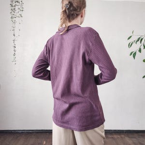 May include: A long-sleeved, ribbed knit shirt in a muted purple hue. The shirt features a collared neckline and a relaxed fit. The fabric has vertical ribbing, adding texture to the garment. The model is wearing beige pants.