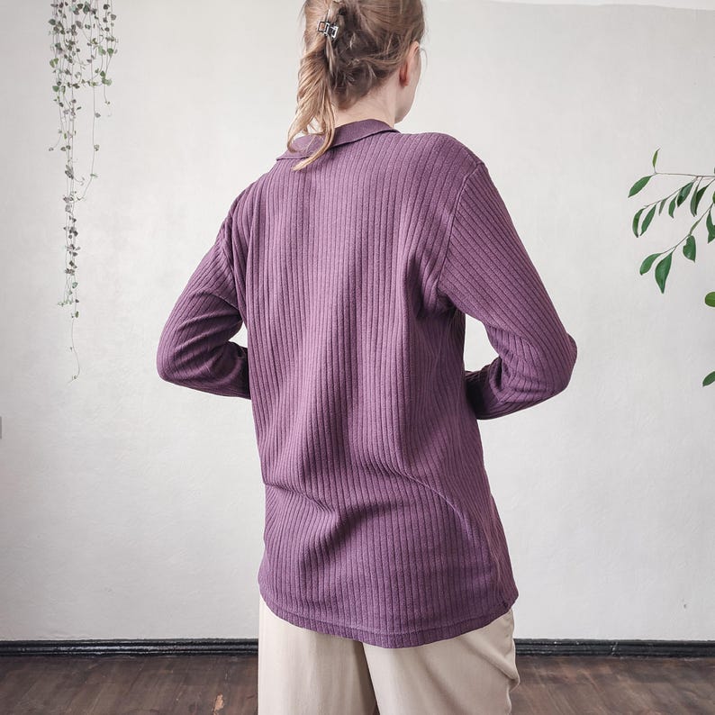May include: A long-sleeved, ribbed knit shirt in a muted purple hue. The shirt features a collared neckline and a relaxed fit. The fabric has vertical ribbing, adding texture to the garment. The model is wearing beige pants.