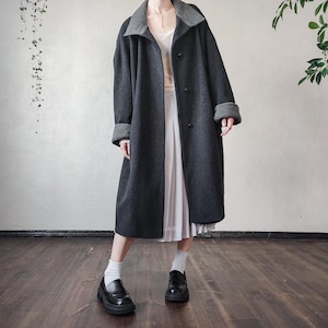 May include: A long, dark gray coat with a contrasting gray collar and cuffs. The coat has a button-down front and a knee-length hem. The coat is paired with a white skirt and black loafers.
