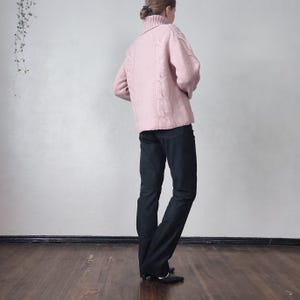 May include: A pale pink cable-knit roll-neck sweater with long sleeves. The sweater is worn with black trousers and black shoes. The outfit is worn by a person standing on a wooden floor.