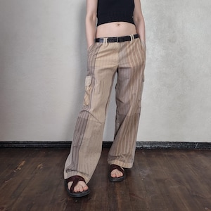 Può includere: Pantaloni cargo beige e marroni con un design a righe e patchwork. I pantaloni hanno due tasche cargo e una cintura nera. La persona indossa sandali marroni.