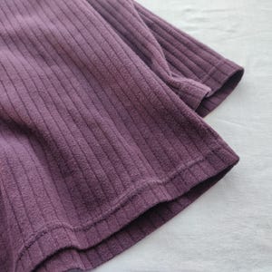 May include: Close-up of a plum-colored ribbed knit fabric. The fabric has a textured surface with vertical lines. The edges of the fabric are visible, showing the hem and sleeve of a garment. The fabric is soft and appears to be made of a comfortable material.