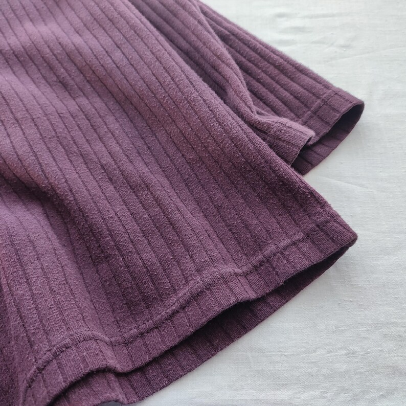 May include: Close-up of a plum-colored ribbed knit fabric. The fabric has a textured surface with vertical lines. The edges of the fabric are visible, showing the hem and sleeve of a garment. The fabric is soft and appears to be made of a comfortable material.