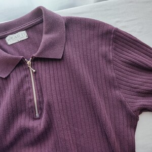 May include: A plum-colored ribbed knit shirt with a collared neckline and a silver zipper. The shirt has short sleeves and the tag reads "WEAR IT". The fabric has a vertical ribbed texture.