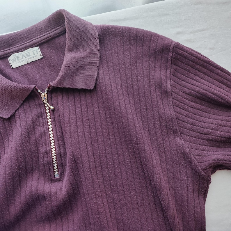May include: A plum-colored ribbed knit shirt with a collared neckline and a silver zipper. The shirt has short sleeves and the tag reads "WEAR IT". The fabric has a vertical ribbed texture.