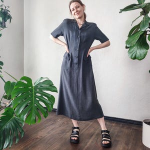 May include: A dark gray midi dress with short sleeves, a button-down front, and two front pockets. The dress is paired with black sandals with white accents. The background features green plants and a wooden floor.