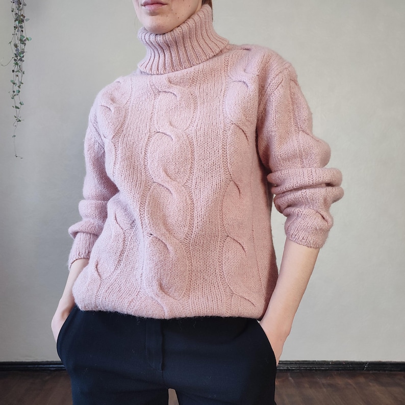 May include: A pale pink cable-knit turtleneck jumper. The jumper has a textured cable design across the front and sleeves. The model is wearing black trousers. The jumper has a high, ribbed turtleneck collar and long sleeves.