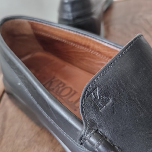 K&ouml;nnte beinhalten: Nahaufnahme eines schwarzen Lederloafers. Der Innenraum ist beige mit dem Markennamen "KROLL" sichtbar. Ein Logo ist auf der Seite gepr&auml;gt. Der Schuh ist auf Komfort und Stil ausgelegt.