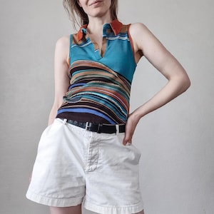 Può includere: Un top senza maniche turchese e a righe con colletto marrone e cintura nera. Il top presenta uno scollo a V ed è abbinato a pantaloncini bianchi. Il top ha strisce orizzontali di vari colori.
