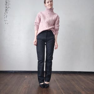 May include: A woman in a light pink cable-knit turtleneck jumper and dark denim jeans. The jeans are straight-legged and worn with black heels. The outfit is a classic, simple style.