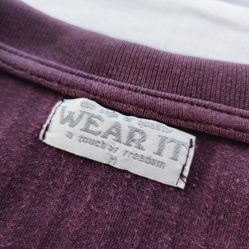 May include: Close-up of a dark purple t-shirt with a ribbed neckline. A rectangular tag is sewn onto the shirt, displaying the words "WEAR IT" in a faded, textured font. The tag also includes the phrases "the sign of quality" and "a touch of freedom".