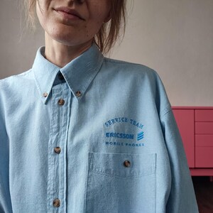 May include: A light blue button-down shirt with a collar and a single chest pocket. The shirt features embroidered text in blue that reads SERVICE TEAM ERICSSON MOBILE PHONES, accompanied by a three-line logo above the pocket.