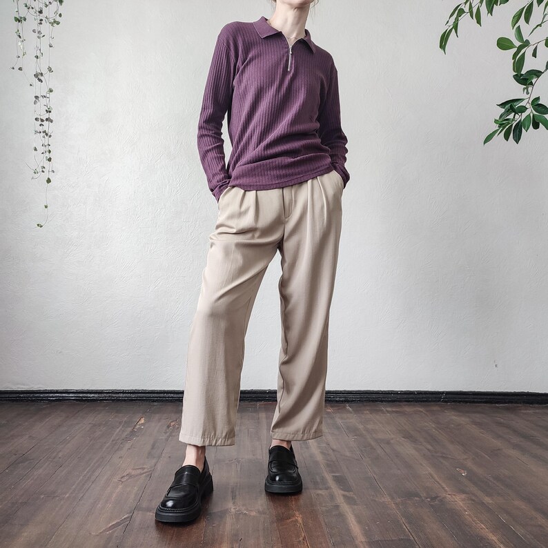 May include: A person wearing a plum-colored, long-sleeved polo shirt with a ribbed texture and a quarter-zip closure. They are wearing beige, wide-leg trousers and black leather loafers. The outfit is styled in a minimalist aesthetic.