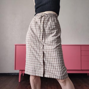 Peut inclure: Une jupe longueur genou avec un motif à carreaux marron et blanc, une taille élastique avec un cordon de serrage et une rangée verticale de boutons sur le devant. La jupe présente deux poches avant et une coupe droite et structurée.