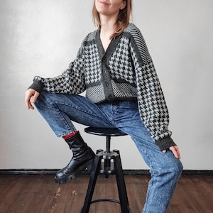 May include: A houndstooth pattern cardigan in shades of gray and black, paired with blue jeans. The cardigan has a V-neck and button closure. The outfit is completed with black boots.