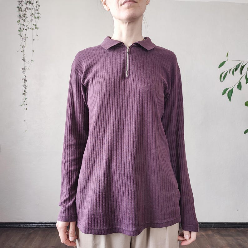May include: A long-sleeved, ribbed knit shirt in a deep purple hue. The shirt features a collared neckline and a silver zipper that extends down the chest. The fabric has vertical ribbing, and the sleeves are long.