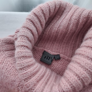 May include: A close-up shot of a dusty rose turtleneck sweater. The sweater features a ribbed knit design on the collar and body. A black label with the text "ril's" is sewn on the inside of the collar, with a size "L" tag.