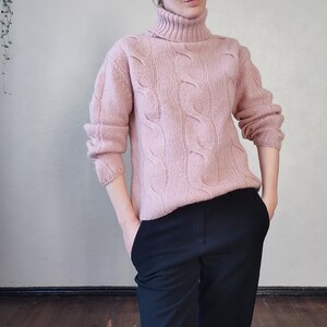 May include: A pale pink cable-knit turtleneck jumper with long sleeves. The jumper features a textured pattern and a high, folded collar. The person is wearing black trousers. The jumper is made of a soft, warm material.