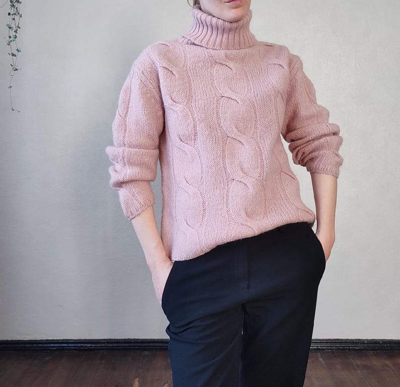 May include: A pale pink cable-knit turtleneck jumper with long sleeves. The jumper features a textured pattern and a high, folded collar. The person is wearing black trousers. The jumper is made of a soft, warm material.