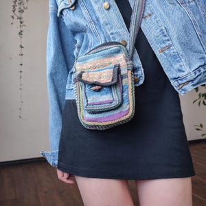 May include: A small, colorful crossbody bag with a woven, striped design. The bag features a front pocket with a wooden toggle closure and a matching striped strap. The color palette includes blue, purple, green, and tan.
