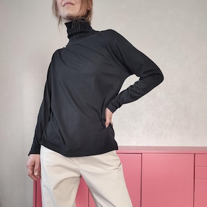 Op de afbeelding: Een effen zwart shirt met lange mouwen en een hoge col. Het kledingstuk wordt gedragen met een lichtbeige broek. Het shirt heeft een eenvoudig, effen ontwerp met lange mouwen en een aansluitende hals voor een strakke en basis uitstraling.