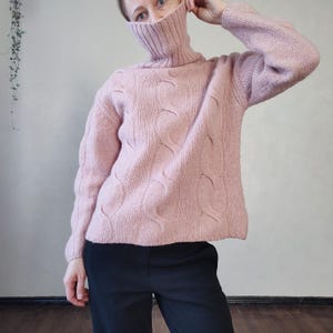 May include: A pale pink cable-knit turtleneck jumper. The jumper features a textured pattern and long sleeves. The model is wearing black trousers. The jumper is made of a soft, warm material.