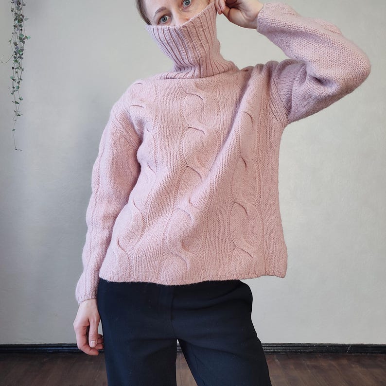 May include: A pale pink cable-knit turtleneck jumper. The jumper features a textured pattern and long sleeves. The model is wearing black trousers. The jumper is made of a soft, warm material.