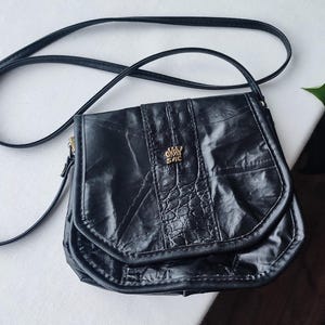 May include: Black crossbody bag with a long strap. The bag features a flap closure and a textured, crocodile-like pattern on the front. The bag has the text "OKAY SAC" in gold lettering.