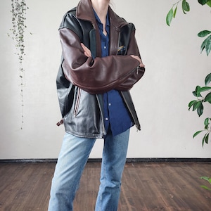 Może przedstawiać: Czarna i brązowa skórzana kurtka z brązowym kołnierzem i rękawami. Kurtka ma zapięcie na zamek błyskawiczny i małą naszywkę na górnej lewej piersi. Osoba ma na sobie niebieską koszulę zapinaną na guziki i niebieskie dżinsy.