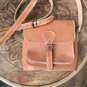 以下が含まれることがあります： フラップ開閉と調節可能なストラップが付いた、タン色の革製クロスボディバッグ。 バッグは白いステッチと金属製のバックルが特徴です。 バッグは木製の表面に置かれています。