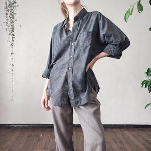 Può includere: Camicia grigio scuro a quadri con un piccolo logo rosa sul petto. La camicia ha maniche lunghe e viene abbinata a pantaloni marrone chiaro. Stile classico.
