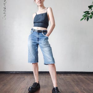 May include: A denim blue crop top with spaghetti straps and light blue denim Bermuda shorts with white trim and a frayed hem. The shorts are worn with a black belt and black platform shoes. The outfit is casual and stylish.