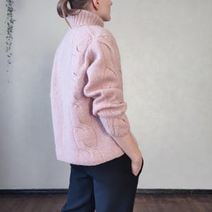 May include: A pale pink cable-knit turtleneck jumper. The jumper features a high, ribbed collar and long sleeves. The cable knit pattern is visible across the front and sleeves. The person is wearing black trousers.