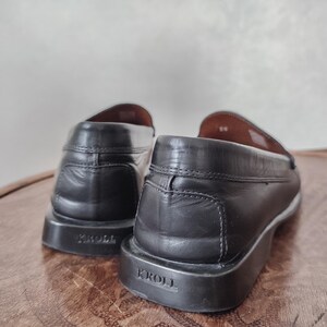 K&ouml;nnte beinhalten: Ein Paar schwarze Leder-Loafer mit dem Markennamen "KROLL" auf der Ferse. Die Schuhe haben ein klassisches Design mit runder Spitze und niedrigem Absatz. Die Innenseite ist hellbraun.