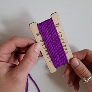 May include: A wooden yarn wrap gauge with purple yarn wound around it. The gauge has a list of yarn weights and corresponding needle sizes printed on it. The yarn weights listed are Super Bulky, Bulky, Chunky, Aran, DK, 4 Ply, Fingering, Lace.