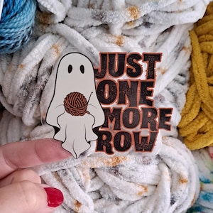 May include: A clear sticker with a cartoon ghost knitting with a ball of yarn, and the text "JUST ONE MORE ROW" in orange and black. The sticker is set against a backdrop of white, blue, and yellow yarn.