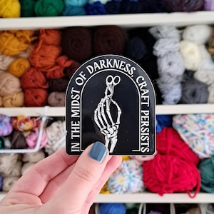 May include: A black sticker with white text and a skeleton hand holding scissors. The text reads "IN THE MIDST OF DARKNESS, CRAFT PERSISTS." The background shows shelves filled with colourful yarn.