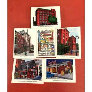 May include: Five vintage postcards depicting different buildings and scenes in Manchester, New Hampshire. The postcards feature illustrations of a cigar factory, a street with shops, a pub, a restaurant, and a corner with a building and a traffic light.