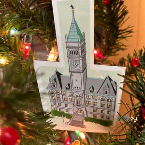 May include: A Christmas tree ornament featuring a detailed illustration of a building with a clock tower. The ornament is made of wood and has a rustic, vintage aesthetic.