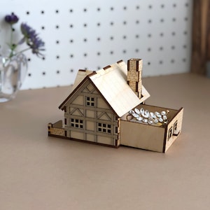 Puede incluir: Una caja de joyer&iacute;a de madera con forma de casa con un caj&oacute;n abierto, que revela un peine de pelo plateado brillante en su interior. La caja est&aacute; hecha de madera clara y tiene un techo y una chimenea marrones.