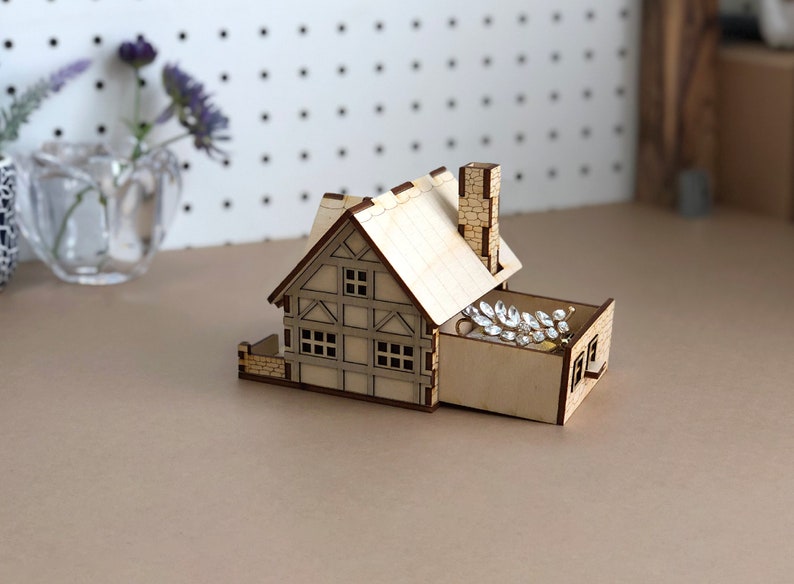 Puede incluir: Una caja de joyer&iacute;a de madera con forma de casa con un caj&oacute;n abierto, que revela un peine de pelo plateado brillante en su interior. La caja est&aacute; hecha de madera clara y tiene un techo y una chimenea marrones.