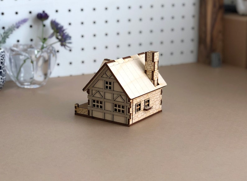 Puede incluir: Un peque&ntilde;o modelo de casa de madera con una chimenea de piedra y ventanas. La casa est&aacute; pintada de color marr&oacute;n claro y tiene un techo blanco.