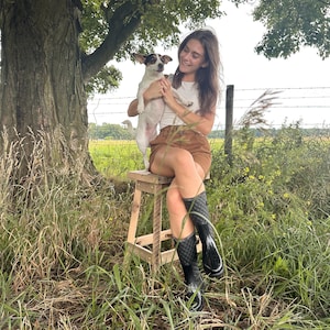 May include: A woman wearing a white shirt, brown skirt, and black rain boots sits on a wooden stool in a field. She is holding a small white and brown dog in her arms.