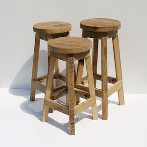 May include: Three wooden stools with round seats and four straight legs. Each stool is constructed from light brown wood with a visible grain pattern and features horizontal support bars connecting the legs for stability and structural reinforcement.