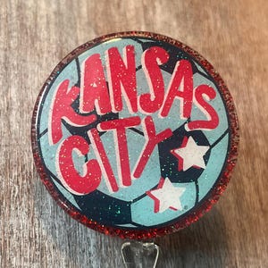 May include: A round badge reel with a red glitter border. The badge reel features the words "Kansas City" in red over a light blue soccer ball design with white stars. A silver clip is attached.
