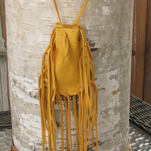 May include: Mustard yellow leather pouch with long fringe. The pouch is suspended by two thin leather straps. The background is a birch tree trunk with a light-colored bark. The pouch is likely used for carrying small items.