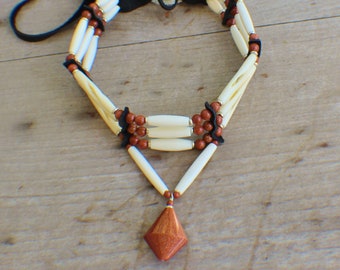 May include: A handcrafted necklace featuring off-white tubular beads, interspersed with reddish-brown beads and black leather accents. A diamond-shaped pendant hangs from the bottom. The necklace is displayed on a wooden surface.