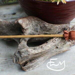 May include: A wooden hair stick with a brown leather accent. The hair accessory is set against a weathered wood background, with a brown pot and greenery in the background. The hair stick is a natural color.